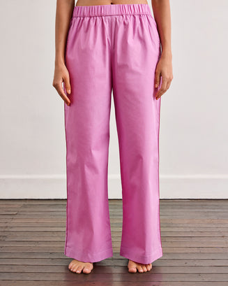 A person stands barefoot on a wooden floor, wearing Bed Threads' Bubblegum & Berry Piped Pants—relaxed, long-sleeved, button-up pink pajamas with an elasticated waist—in front of a plain white wall.