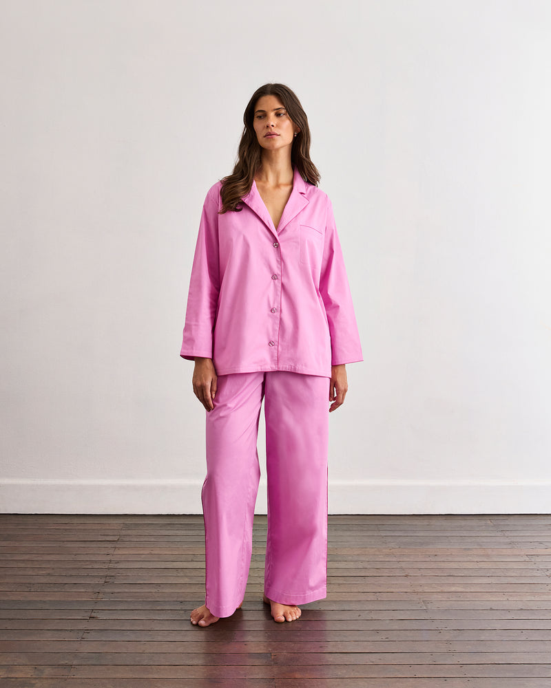 A person stands barefoot on a wooden floor, wearing loose high-waisted pink pants from the Bed Threads Bubblegum & Berry Piped Long Sleep Set; the elastic waistband is visible, but the upper body is not shown.