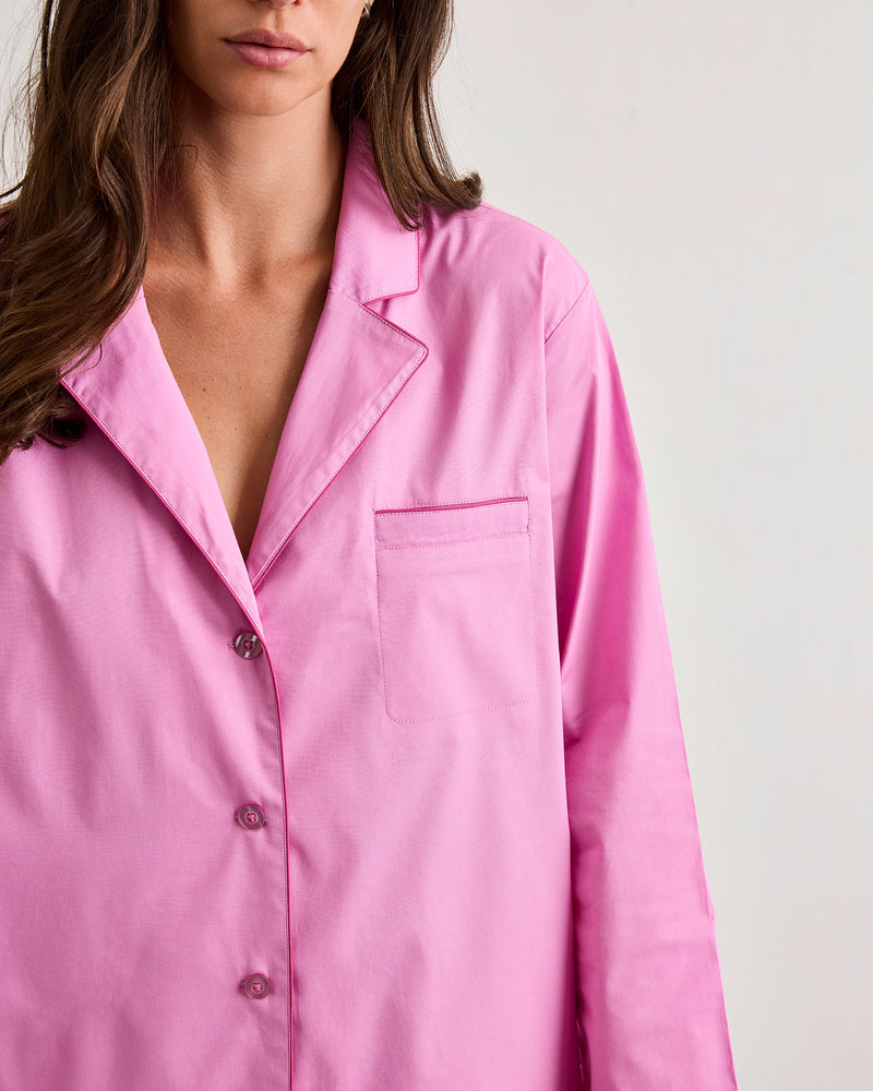 A woman wears the Bed Threads Bubblegum & Berry Piped Long Sleeve Shirt, a loose-fitting, bright pink cotton pyjama-style button-up with a chest pocket, standing against a plain light background. Her face is partially out of frame.