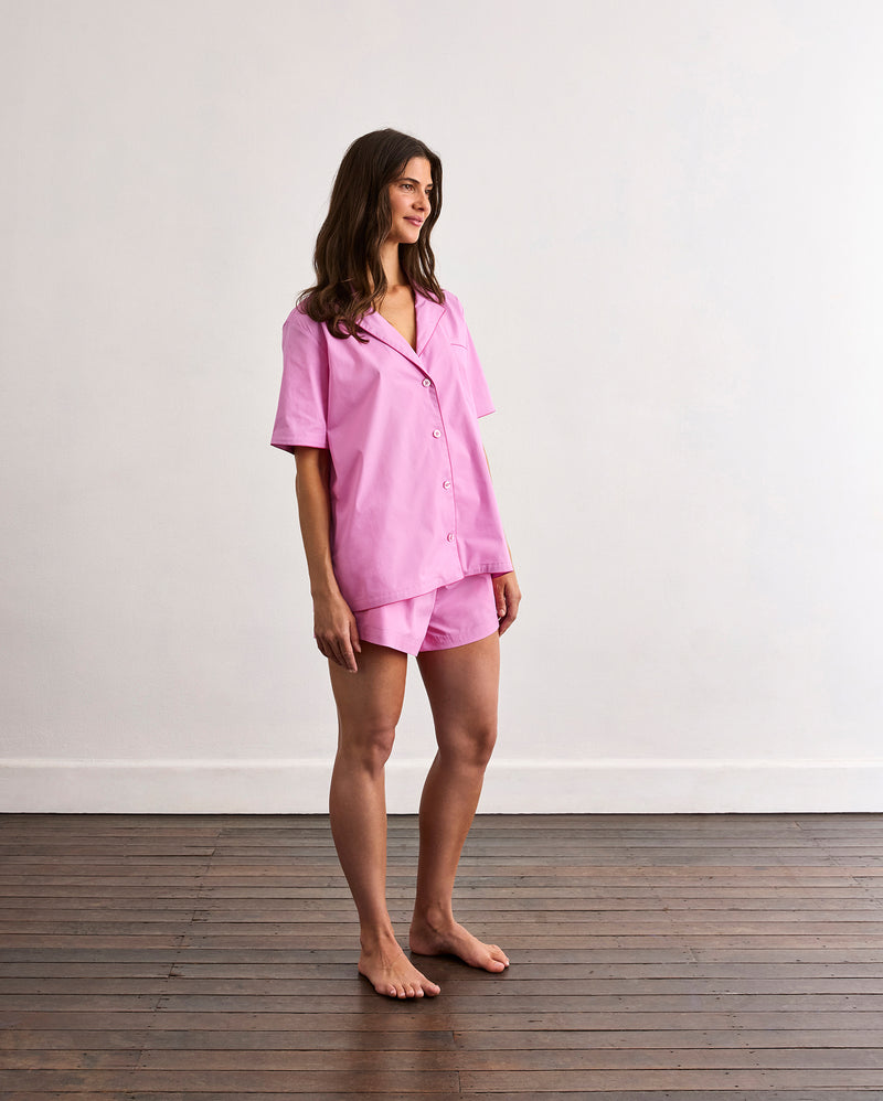 A woman stands barefoot on a wooden floor against a white wall, wearing Bed Threads' Bubblegum & Berry Piped Shorts sleepwear. She faces slightly to the side and smiles gently, showcasing the short-sleeved, loose cotton set.