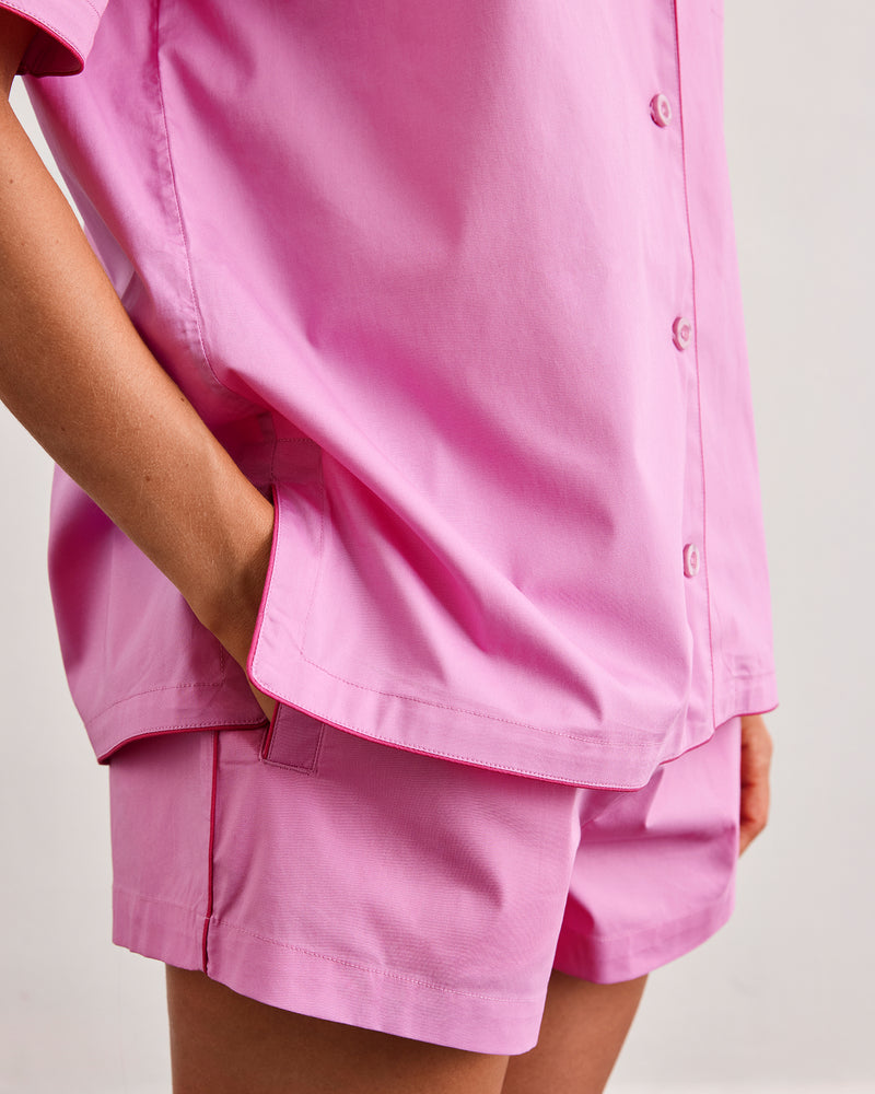 A person models the Bed Threads Bubblegum & Berry Piped Shorts—a loose-fitting, short-sleeved pink button-up shirt and matching piped shorts—standing with one hand in their pocket against a plain background.