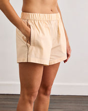A person wears Bed Threads Vanilla & Espresso Piped Shorts—light beige cotton with an elastic waistband and pockets—standing against a plain white wall and wooden floor, shown from midriff to mid-thigh. - view