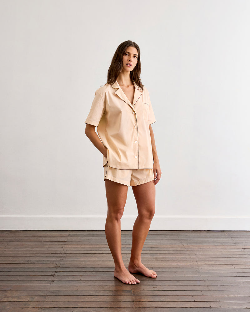 A woman stands barefoot on a wooden floor against a plain white wall, wearing Bed Threads' Vanilla & Espresso Piped Shorts—a short-sleeve, button-up beige pajama shirt and matching breathable shorts.