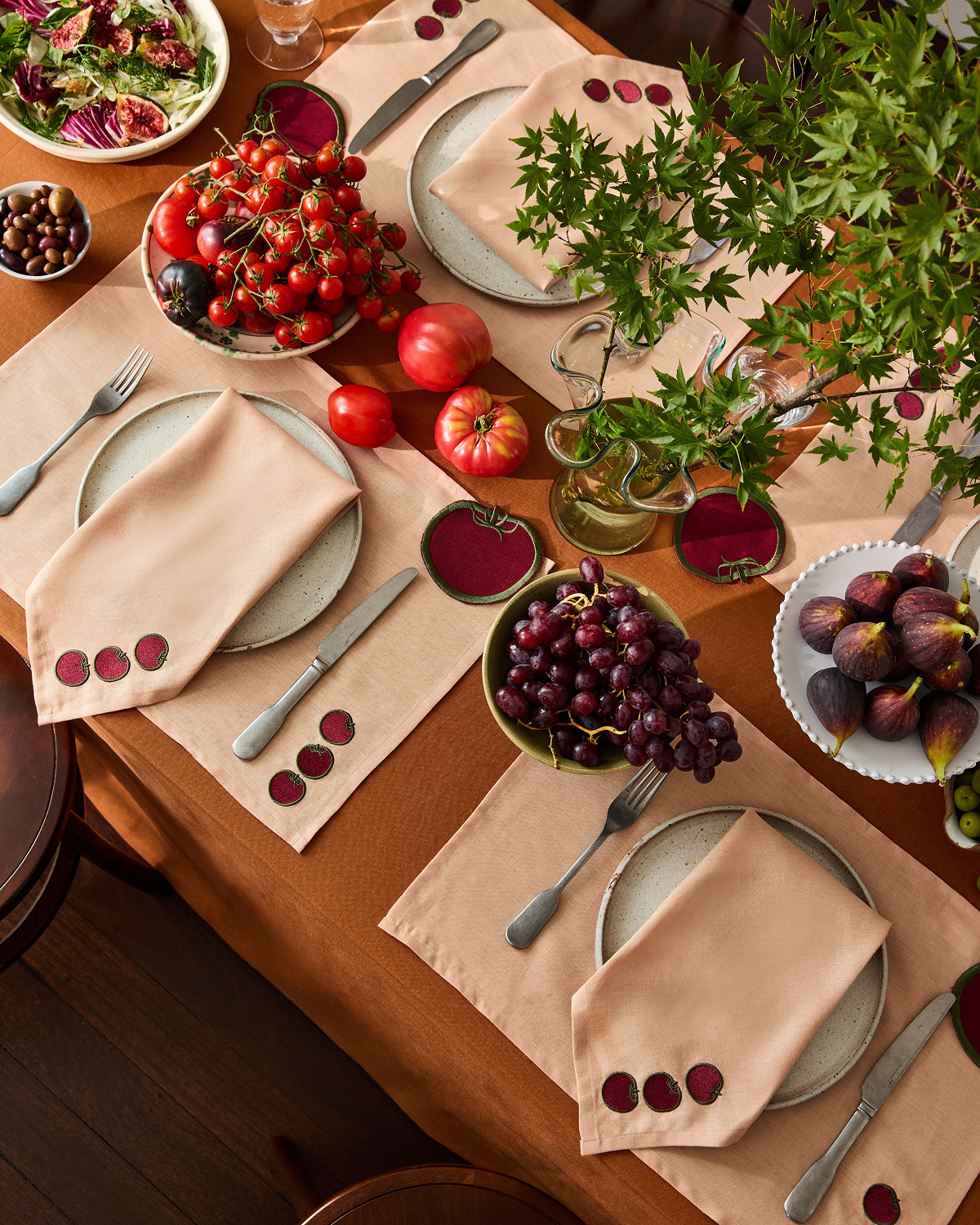 A table set with beige napkins, placemats, plates, cutlery, and glasses is accented by Bed Threads' Les Fruits Tomate Coasters (Set of Four), fresh tomatoes, grapes, figs, a green leafy branch, bowls of olives, and salad.