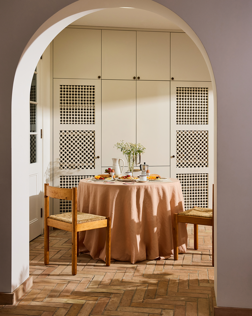 A round table draped in the Bed Threads Terracotta 100% French Flax Linen Tablecloth is set for breakfast with plates, cups, and flowers, flanked by two wooden chairs in a sunlit room with white cabinets and terracotta tile floors.