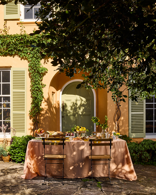 A Terracotta 100% French Flax Linen Tablecloth by Bed Threads dresses an outdoor table set for dining in front of a yellow house with green shutters, surrounded by plants and sunlight for a luxurious setting.