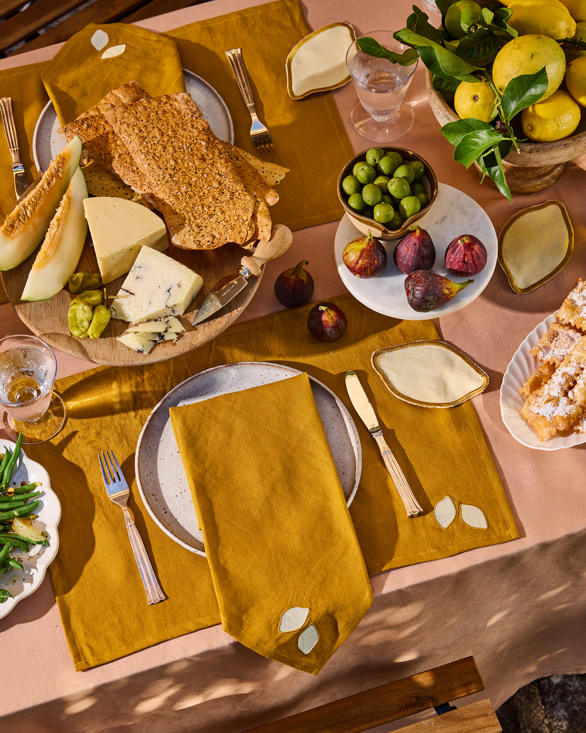 A luxury table setting showcases mustard yellow plates, cheese, crispbread, melon, figs, green olives, pastries, and lemons with leaves atop the Terracotta 100% French Flax Linen Tablecloth by Bed Threads.