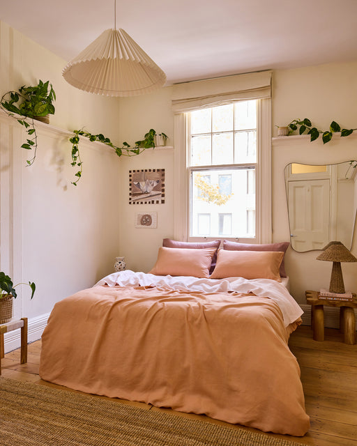 A cozy bedroom with Bed Threads’ Rosewater 100% French Flax Linen Sheet Set, a window above the bed, plants on shelves, a rattan rug, a pendant light, a side table with a woven lamp, and neutral-toned walls and decor.