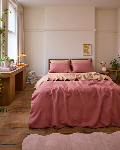 A cozy bedroom features a Bed Threads Pink Clay 100% French Flax Linen Bedding Set, a wooden headboard, wooden floors, a soft pink rug, a desk with plants and books, all bathed in natural light from a large window.