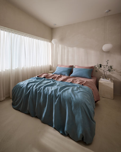 Minimalist bedroom with a large bed dressed in blue and pink bedding, Bed Threads’ Lavender 100% French Flax Linen Pillowcases, sheer white curtains, white pendant lights, and a small plant on a marble nightstand.