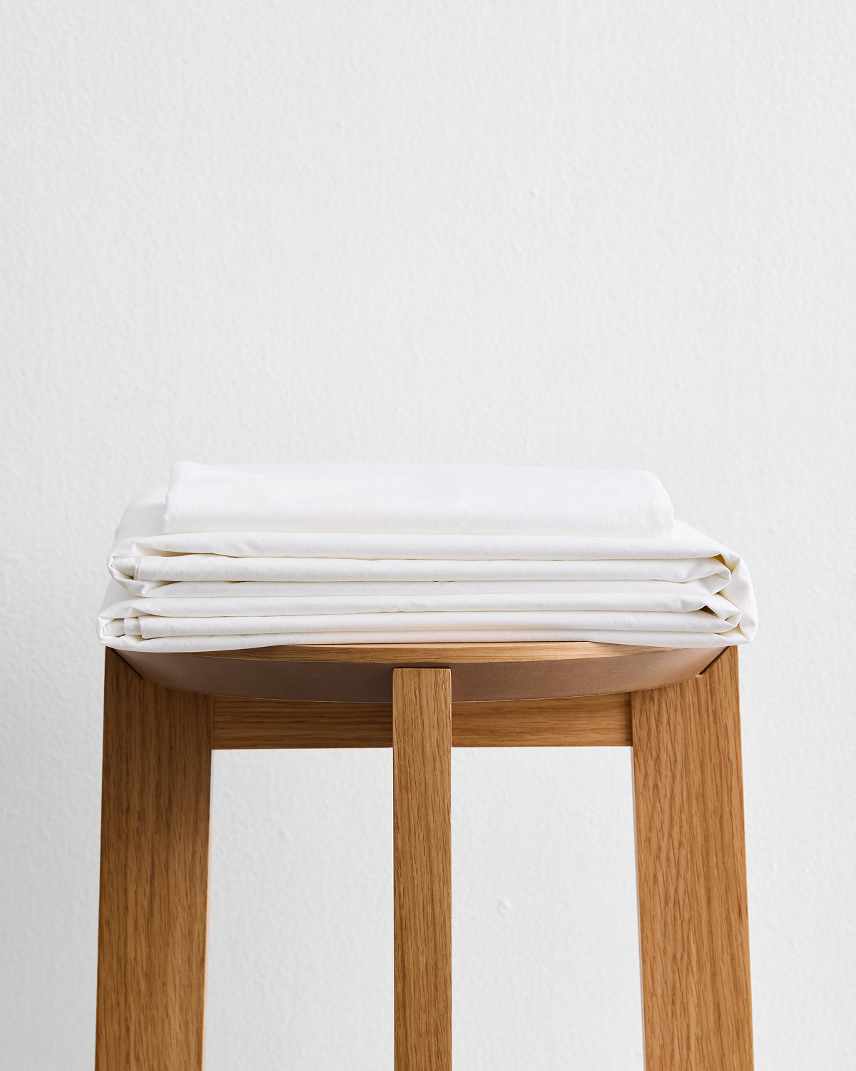A neatly folded stack of Bed Threads Crisp White Organic Cotton Sheet Set rests on a light wooden stool against a plain white wall.