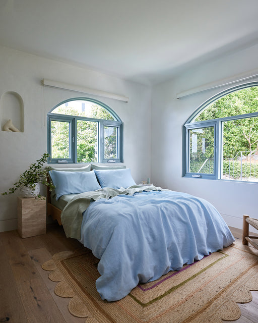 A bright bedroom features the Sage 100% French Flax Linen Fitted Sheet by Bed Threads, wooden floors, a woven rug, two large arched windows with natural light, and a small bedside table holding a vase of greenery.