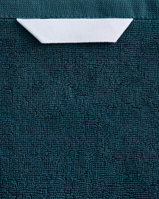 A close-up of the Bed Threads Aegean Cotton Terry Bath Towel Bundle in dark teal, featuring a white rectangular paper tab and showcasing its highly absorbent textured material.