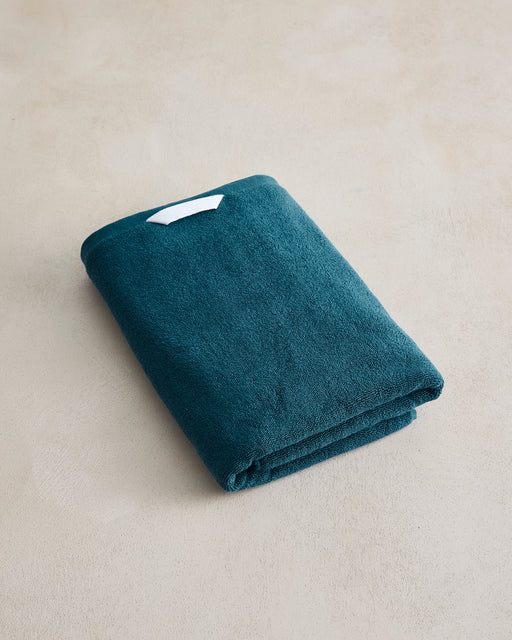 A neatly folded Aegean Cotton Terry Bath Towel by Bed Threads in dark teal, featuring a small white label on top and placed on a light beige surface.
