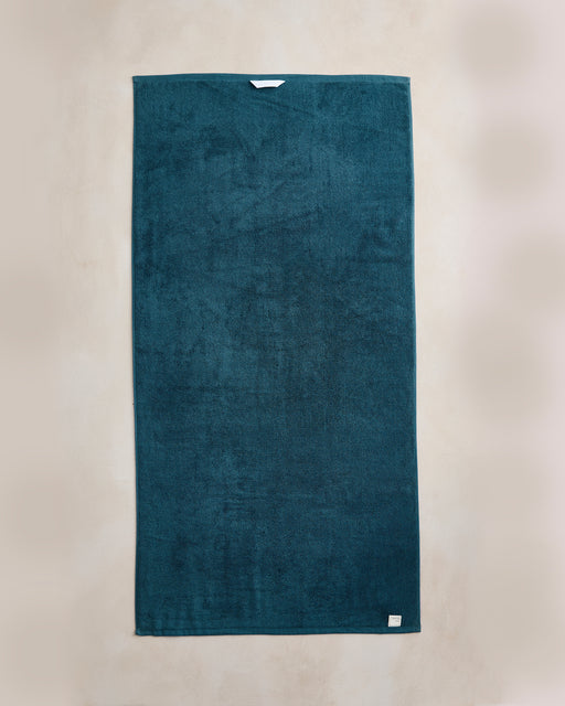 The Bed Threads Aegean Cotton Terry Bath Towel is neatly laid out in a rectangle, teal in color with a small white label at the bottom right, displayed on a light beige background.