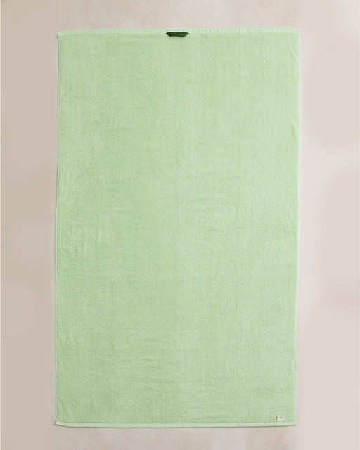 The Bed Threads Apple Cotton Terry Bath Sheet is a rectangular, light green towel made from plush quick-drying cotton, featuring a small corner tag and a hanging loop near the top edge.