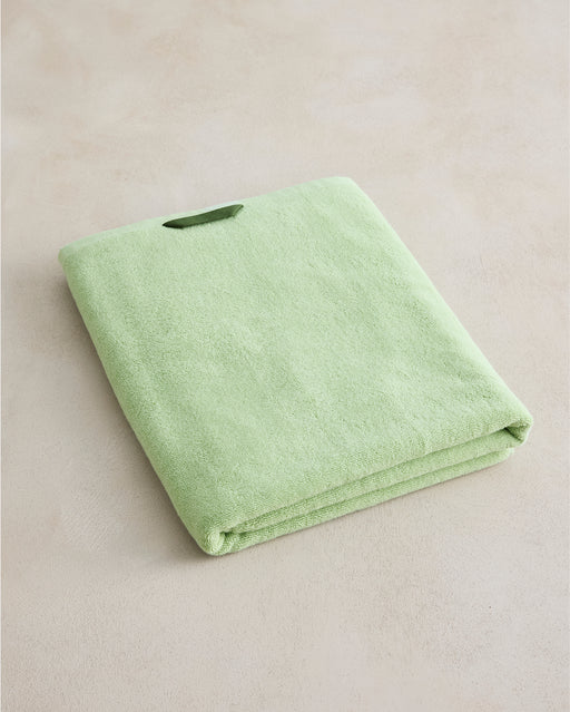 The Bed Threads Apple Cotton Terry Bath Sheet, in light green, is neatly folded with a small loop tag and rests on a beige surface.