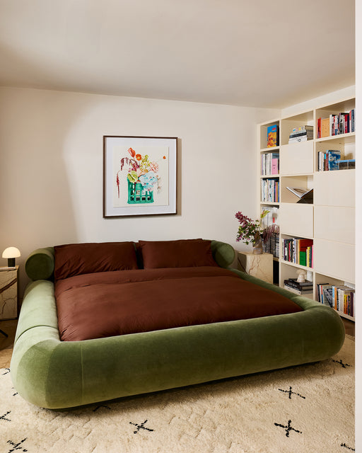 A cozy bedroom with a large green upholstered bed, brown bedding, Bed Threads' Espresso Organic Cotton Pillowcases, a beige rug with black patterns, built-in bookshelf, small lamp, and colorful artwork above the bed.