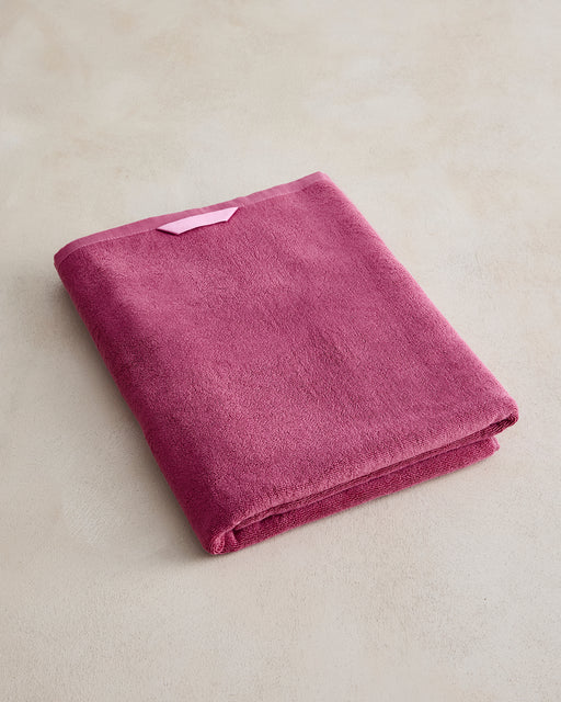 The Berry Cotton Terry Bath Sheet by Bed Threads, in dark pink with a light pink trim and hanging loop, rests on a light beige surface, bringing a luxurious touch to your bathroom.