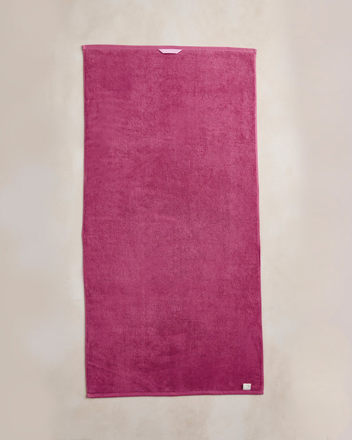 The Berry Cotton Terry Bath Towel from Bed Threads, made of 100% cotton in magenta, is laid flat on a light beige background with a small white tag visible on one edge.