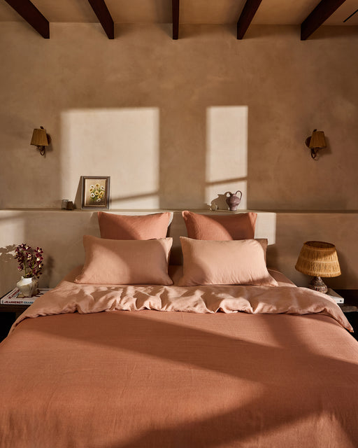 A cozy bedroom features a neatly made bed with peach bedding and the Terracotta 100% French Flax Linen Flat Sheet by Bed Threads. Sunlight casts warm shadows on a beige wall adorned with shelves holding art, a ceramic vase, and decor.