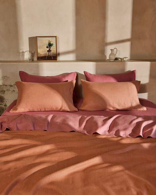 A neatly made bed features Bed Threads’ Pink Clay 100% French Flax Linen Pillowcases (Set of Two), paired with orange and pink bedding, large and small pillows, all set against a beige wall with sunlight. Decorative items and a framed picture sit on a shelf above.