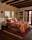 A bed dressed in the Bed Threads Hazelnut & Pink Clay 100% French Flax Linen Bedding Bundle sits against a beige wall, with a shelf above holding decor like a framed painting, glass, white vase, and ceramic pitcher. Shadows fall across the scene.