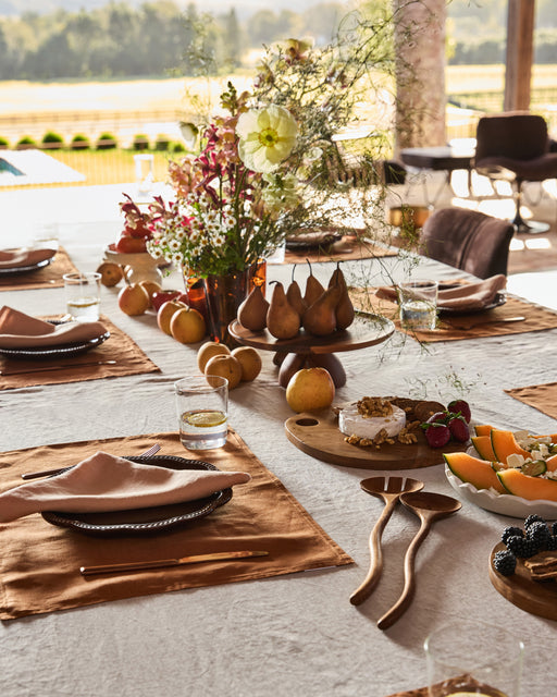 An outdoor dining table with a beige tablecloth, Bed Threads Terracotta 100% French Flax Linen Napkins, glassware, a floral centerpiece, and platters of pears, melon, and berries shines in natural sunlight.