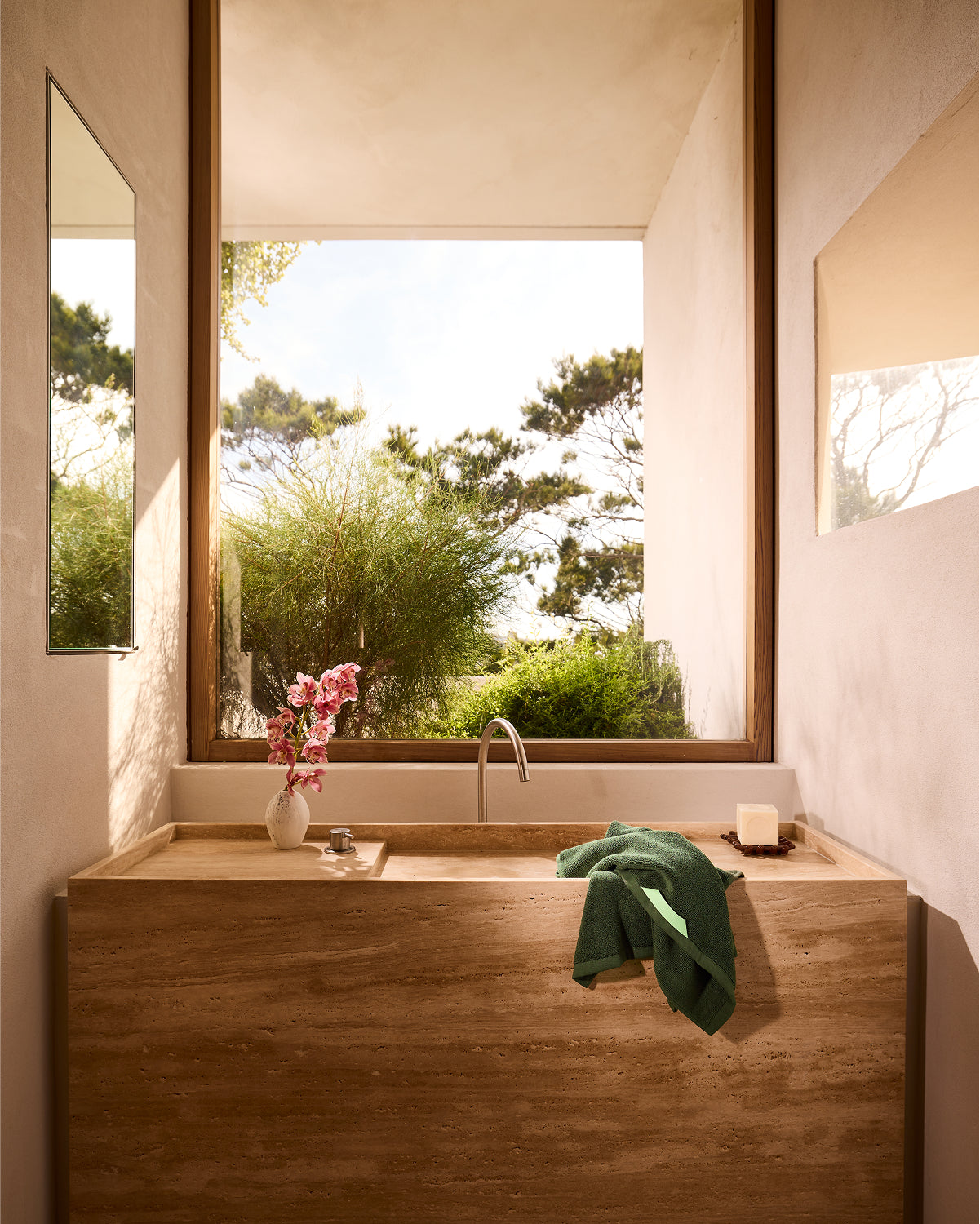 A modern bathroom features a large stone sink, a vase of pink flowers, and a plush Bed Threads Moss Cotton Terry Hand Towel beside soap. Sunlight pours through a big window overlooking lush greenery and trees outside.