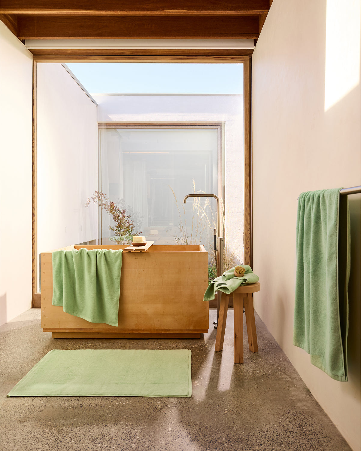 A minimalist bathroom features a wooden tub, Bed Threads’ Apple Cotton Terry Bath Sheet, and a matching bath mat. Sunlight pours through large windows onto neutral walls and a polished concrete floor. A wooden stool holds bath accessories.