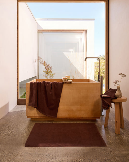 A minimalist bathroom features an elegant wooden bathtub, Bed Threads Espresso Cotton Terry Bath Towels (Set of Two), a matching brown rug, and a small vase with dried flowers on a stool, all bathed in natural light from a large window.