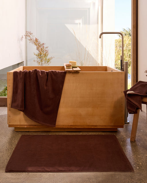 A modern bathroom features an Espresso Cotton Terry Bath Mat by Bed Threads, a wooden soaking tub with brown towels on the tub and bench, and a window offering a garden view in the background.