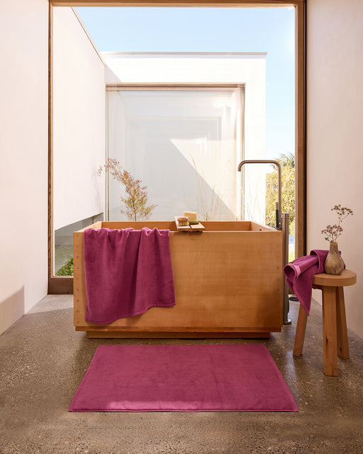 A modern bathroom featuring the Bed Threads Berry Cotton Terry Bath Towel and matching bath mat, plus a wooden bathtub and stool with a vase; a large window fills the space with natural light.