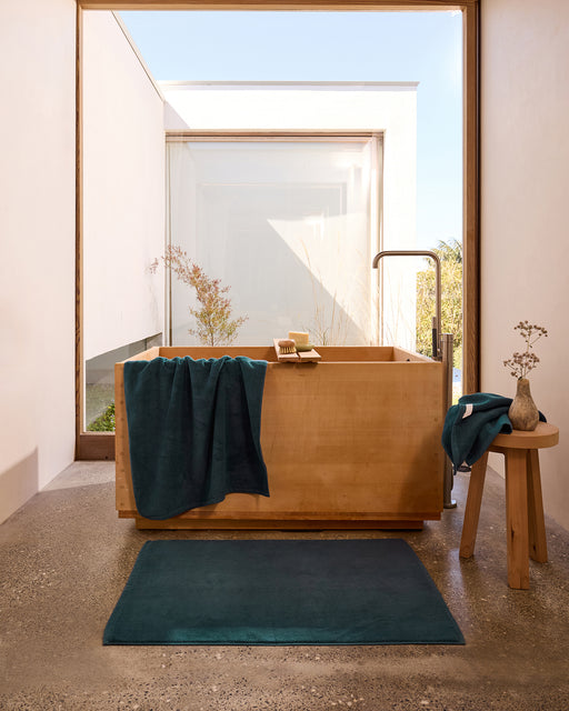 A minimalist bathroom features a wooden soaking tub, a plush Bed Threads Aegean Cotton Terry Bath Towel in teal, a small wooden stool with a vase, and a large window overlooking a serene outdoor garden.