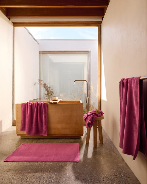 A modern, minimal luxury bathroom with a large wooden tub, floor-to-ceiling window, concrete floor, and plush magenta towels and Bed Threads Berry Cotton Terry Bath Sheets on a stool, rack, and bathmat adding vibrant color.