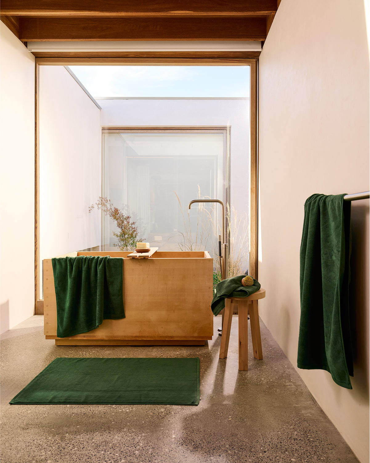 Minimalist bathroom with a wooden soaking tub, small stool holding plush quick-drying towels, a green bath mat, and the Moss Cotton Terry Bath Sheet by Bed Threads hanging on the rack. Natural light highlights the neutral and green decor.