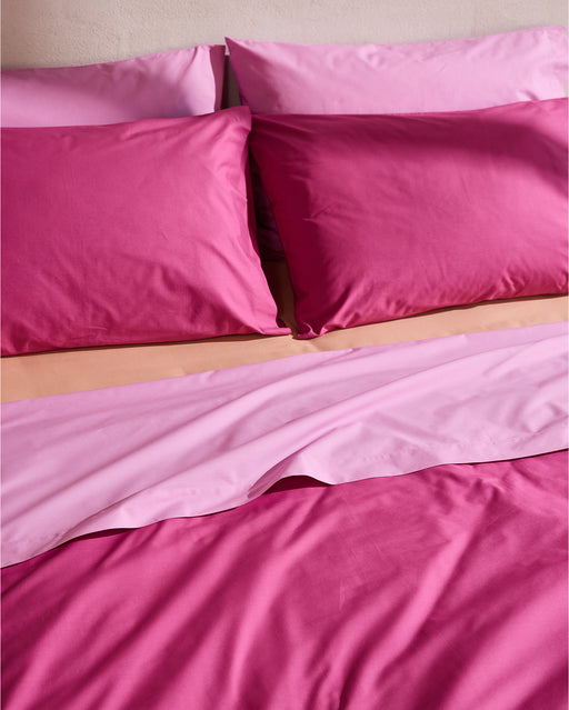 A neatly made bed with the Berry, Bubblegum & Butterscotch Organic Cotton Bedding Bundle by Bed Threads: light pink flat sheet partially turned down over a percale duvet cover and matching pillows, all against a light-colored headboard.