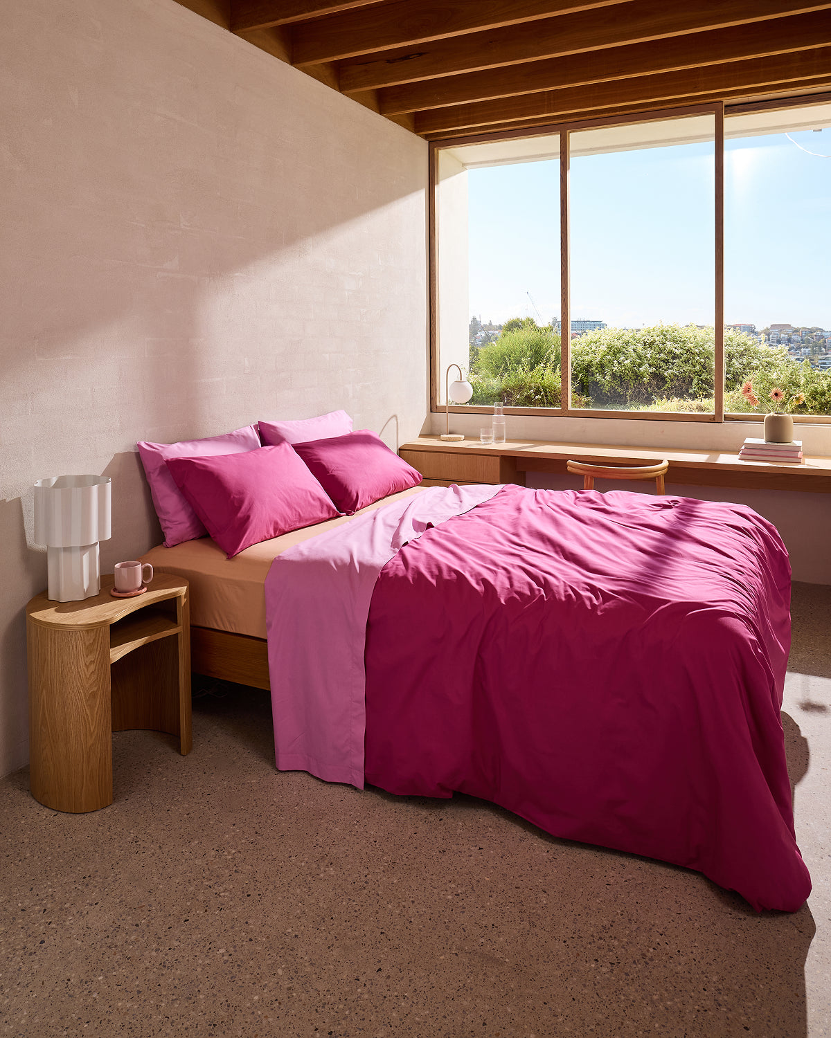 A sunlit bedroom features a bed with vibrant pink and light purple bedding, including the Bed Threads Butterscotch Organic Cotton Fitted Sheet, wooden nightstands, a white lamp, a mug, and a large window overlooking greenery and blue sky.