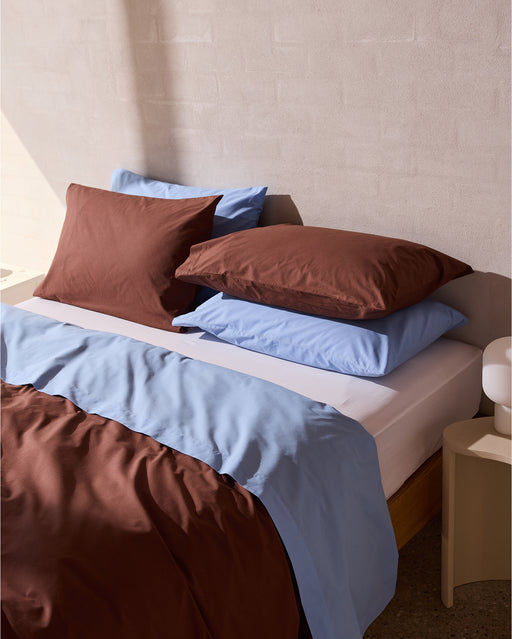 A neatly made bed features the Bed Threads Espresso, Sky & Crisp White Organic Cotton Bedding Bundle—sheets, duvet, and pillowcases—against a cream textured wall. Sunlight casts soft shadows across the luxurious bedding and surrounding areas.