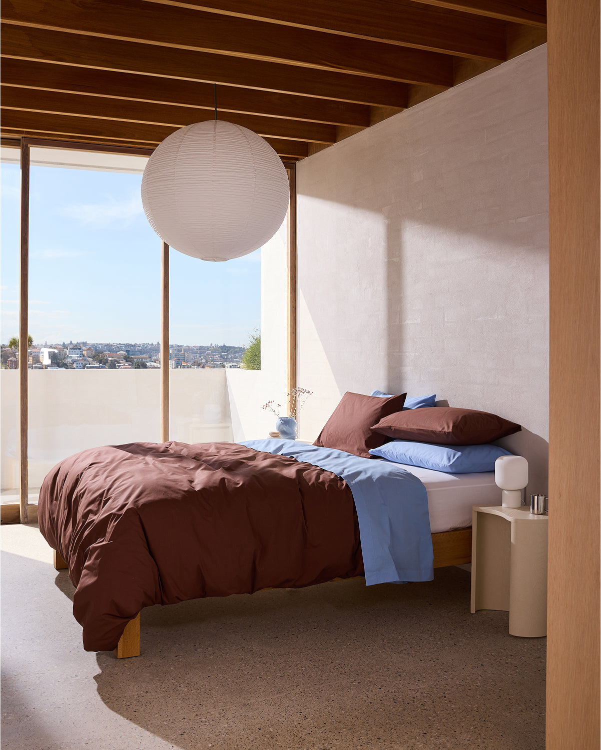 Minimalist bedroom with a large window and wooden ceiling beams features a bed dressed in the Bed Threads Crisp White Organic Cotton Fitted Sheet. A paper lantern hangs above a nightstand with a vase, overlooking the cityscape.
