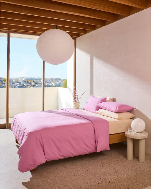 A modern bedroom features a wooden ceiling, large windows with city views, and a bed dressed in the Bed Threads Vanilla Organic Cotton Flat Sheet. A round paper lantern hangs above, while a small side table with a lamp and vase sits nearby.