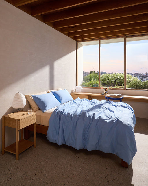 A cozy bedroom features a wooden bed dressed in the Sky Organic Cotton Duvet Cover Set by Bed Threads, with a matching nightstand, lamp, glass of water, large window with views of greenery and buildings, and sunlight streaming in.