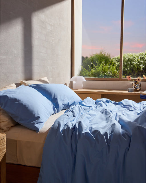 The Bed Threads Sky & Vanilla Organic Cotton Bedding Bundle in light blue adds coziness by the window, where sunlight brightens the room and views of trees and a pink-blue sky create a peaceful, inviting atmosphere.
