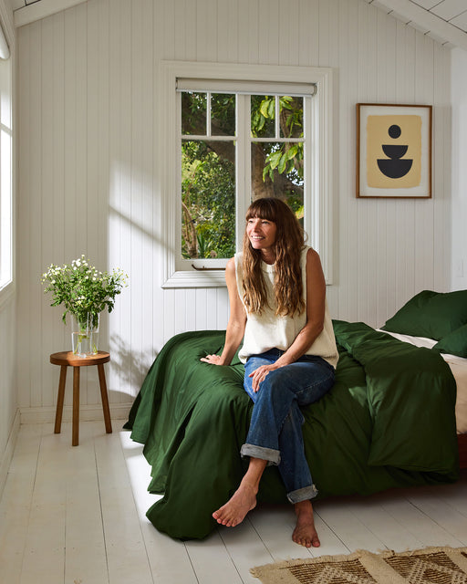 A woman with long brown hair sits barefoot on a bed with green bedding and Moss Organic Cotton King Pillowcases by Bed Threads, smiling as she looks out a bright window. A vase of flowers rests on the bedside table.