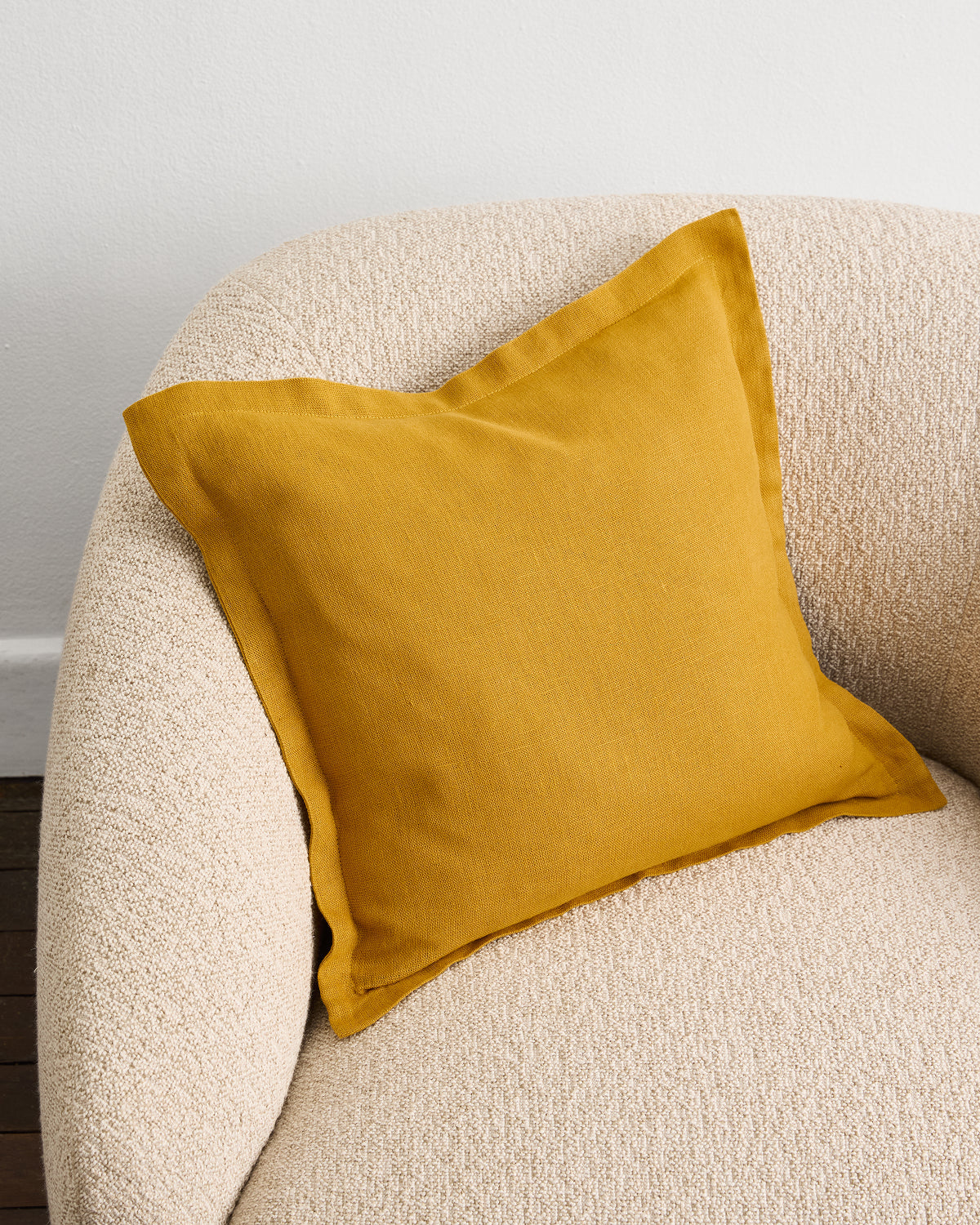 A Bed Threads Turmeric 100% French Flax Linen Cushion Cover in mustard yellow sits on a textured beige armchair by a white wall, with part of a wooden floor visible in the corner.
