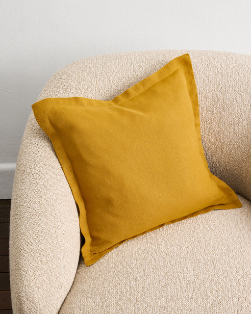 A Bed Threads Turmeric 100% French Flax Linen Cushion Cover in mustard yellow sits on a textured beige armchair by a white wall, with part of a wooden floor visible in the corner.