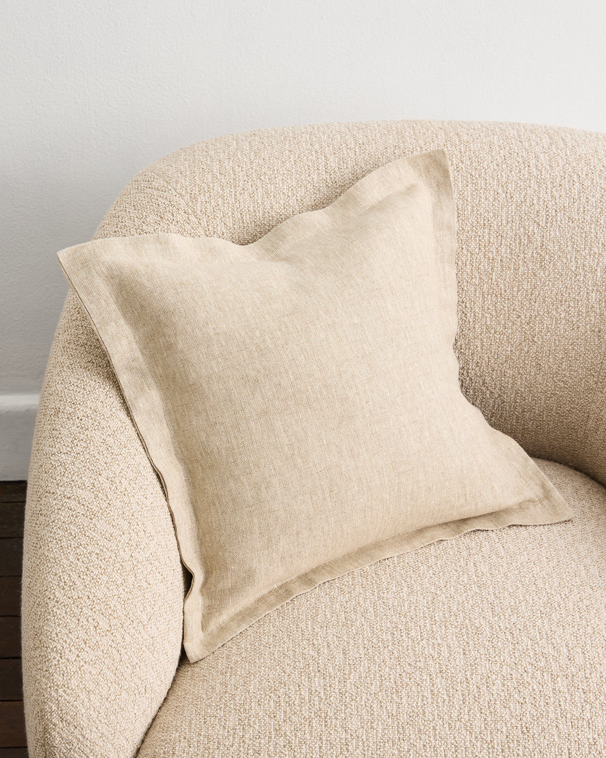 An Oatmeal 100% French Flax Linen Cushion Cover by Bed Threads sits on a textured light beige armchair against a white wall, creating a neutral and cozy atmosphere.