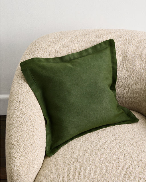 The Olive 100% French Flax Linen Cushion Cover by Bed Threads sits on a textured, light beige armchair with curved edges, set against a plain white wall.