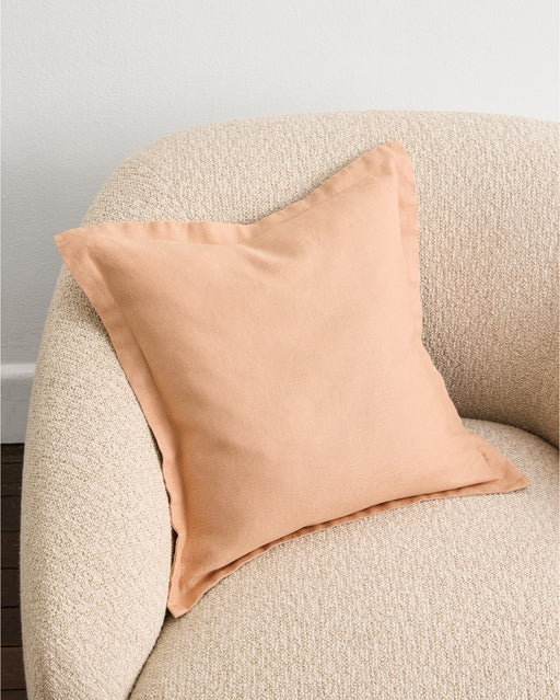 A Terracotta 100% French Flax Linen Cushion Cover by Bed Threads sits on a light beige, textured armchair against a white wall.