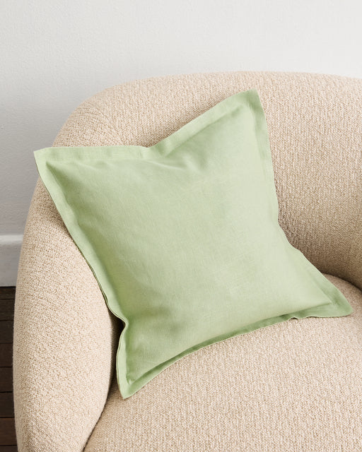The Sage 100% French Flax Linen Cushion Cover by Bed Threads, featuring a flange edge and light green hue, rests on a textured beige armchair near a white wall and wooden floor.